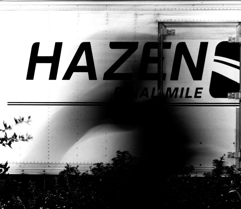 High-contrast black-and-white photograph of an unidentified blurred figure crossing in front of a parked silver Hazen Final Mile truck. The truck’s large lettering remains sharp while the figure dissolves into a dark vertical form.