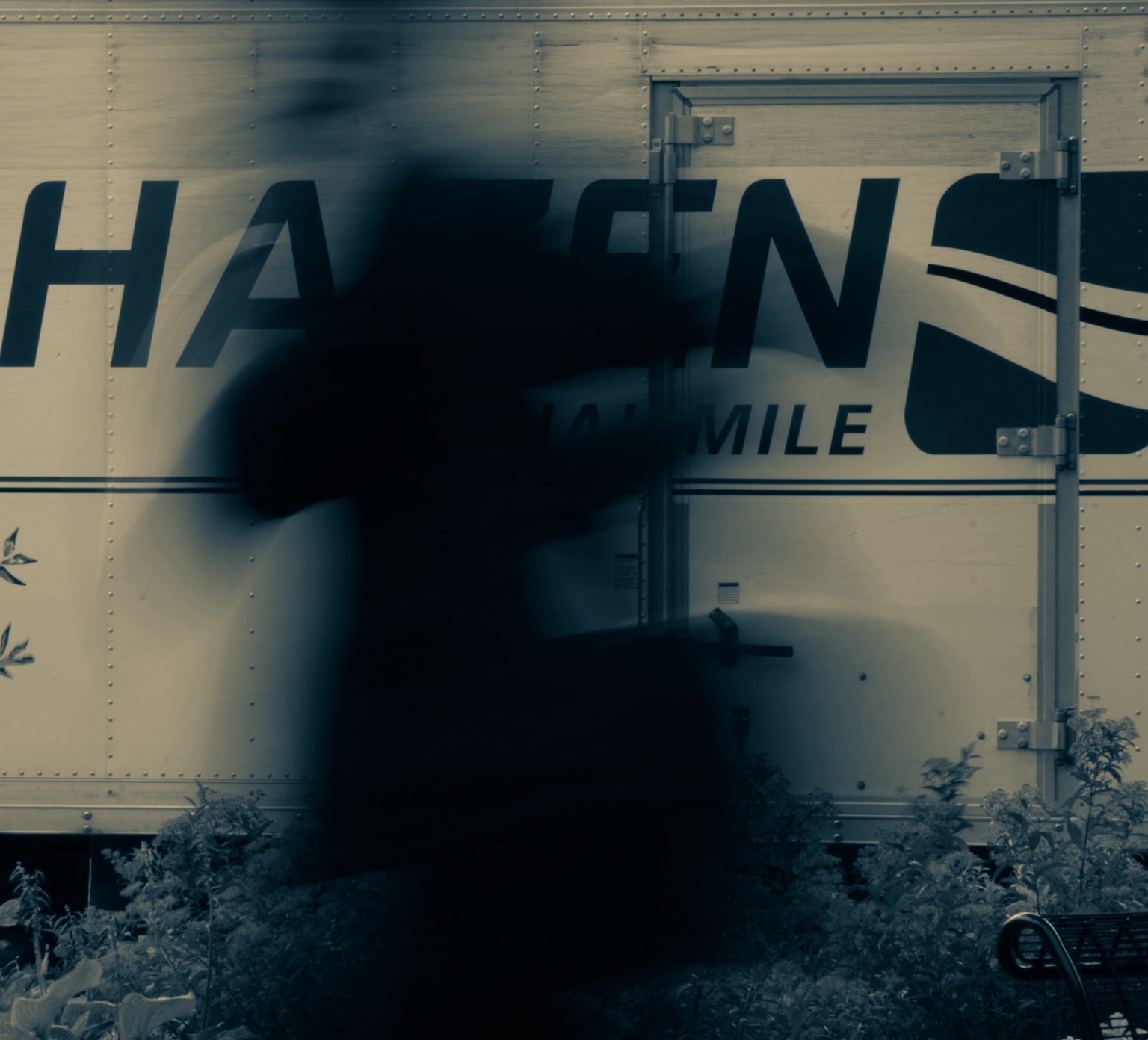 Blue-green-toned photograph of an unidentified blurred figure crossing in front of a parked silver Hazen Final Mile truck. The truck’s lettering and side door remain visible behind the dark moving form.